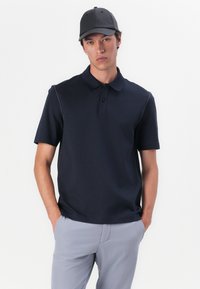 Tumes sinine polo särk lühikeste varrukatega, nööpidega krae ja kontrastsete õmblusdetailidega. ühendatud heledate hallide pükstega. Mütš sama värviga.