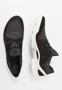 Musta-harmaat tennarit, joissa yhdistyvät sileä ja teksturoitu kangas, valkoinen pohja ja kontrastiset ompelut.