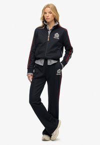 Chándal navy con rayas rojas en los lados, chaqueta con cremallera y logotipo bordado. Camiseta de rayas debajo, combinada con zapatillas de color claro.