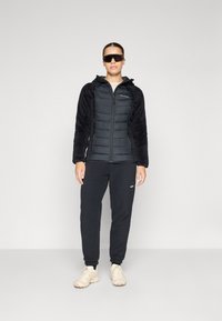 Svart pufferjakke med plysj svarte ermer, glidelås foran og hette. Kombinert med svarte joggebukser og beige sneakers.