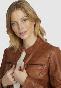 Bruine leren jas met ritszakken, voorzien van een hoge kraag en een gladde textuur. Model houdt de rits vast.