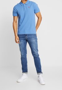 Polo pour homme en coton bleu avec patte de boutonnage à trois boutons et petit logo, associé à un jean en denim et des baskets blanches. Tenue décontractée.