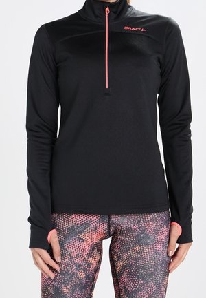 Czarna sportowa bluzka z długim rękawem z różowym zamkiem błyskawicznym i logo, noszona z różowo-czarnymi legginsami we wzory, ręce luźno opuszczone wzdłuż ciała.