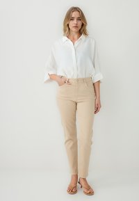 Pantalons en coton beiges au design ajusté, assortis à un chemisier blanc ample avec col et manches retroussées. Sandales marron.