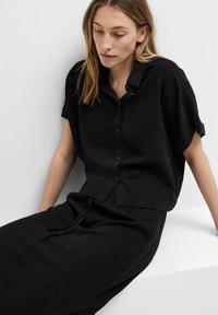 Chemise noire en lin à manches courtes avec un devant boutonné, coupe décontractée et manches retroussées. Associée à un pantalon noir assorti et placée sur un fond neutre.