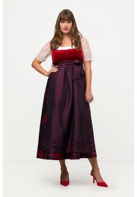 Eine Frau trägt ein traditionelles Kleid mit einem roten Samt-Oberteil, weißen Spitzenärmeln, einem dunkelvioletten Satinrock und roten High Heels und steht vor einem weißen Hintergrund.
