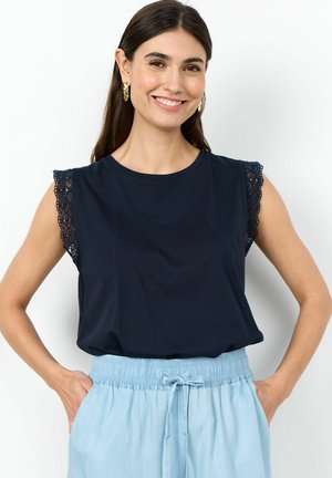 Lächelnde Frau mit langen dunklen Haaren trägt ein marineblaues ärmelloses Oberteil mit Spitzenbesatz und hellblaue, hoch taillierte Hosen mit Kordelzug.