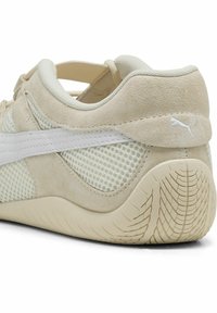 Zapatilla beige claro con parte superior de malla, acentos de gamuza y suela de goma texturizada. Detalle del logo blanco en el talón.