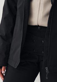 Zwarte buitenjas met ritssluiting, beige fleece voering zichtbaar; zwarte high-waisted broeken met knoopaccenten. Soepele en gestructureerde materialen.