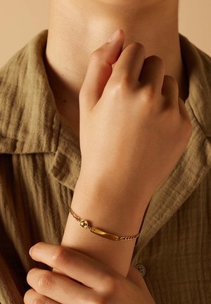 Gouden armband met een gladde staaf en een getextureerde ketting, met een ronde, kraalachtige accent. Draagt om een pols, met een neutrale gekleurde stofachtergrond.