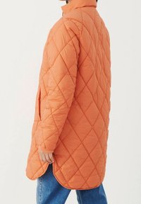 Quiltet orange lang jakke med høj hals, der har sidelommer og ribbede manchetter, vist på en person iført blå jeans.