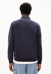 Sweatshirt bleu marine en mélange de coton avec un col et des poignets côtelés, proposant une coupe décontractée. Porté sur une chemise blanche, vu de dos.