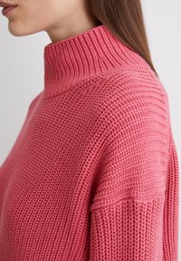 Strickpullover mit hohem Kragen aus einem strukturierten hellrosa Stoff. Verfügt über ein Rippendesign entlang des Kragens und der Schultern.