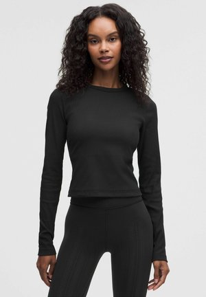 Kvinde med krøllet hår iført en tætsiddende sort langærmet top og teksturerede sorte leggings, stående foran en ensfarvet grå baggrund.