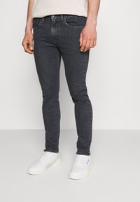 Jeans de mezclilla gris oscuro de corte slim-fit con diseño de cinco bolsillos, sutiles marcas de desgaste y zapatillas bajas blancas. Textura suave con un ligero estiramiento.