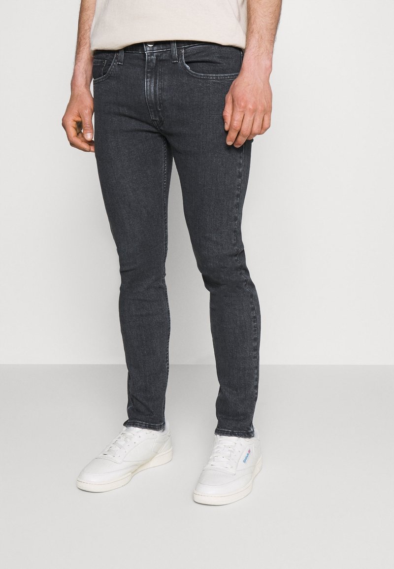 Jeans de mezclilla gris oscuro de corte slim-fit con diseño de cinco bolsillos, sutiles marcas de desgaste y zapatillas bajas blancas. Textura suave con un ligero estiramiento.