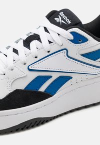 Basket de couleur blanche et bleue en cuir et suède, avec trois bandes bleues, des lacets blancs et un accent noir sur la semelle.