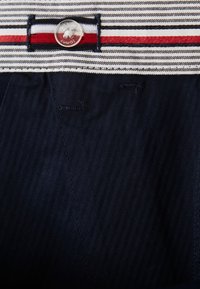 Marina blå byxor med ett randigt, elastiskt midjeband i vitt, rött och marinblått. Inkluderar en silvrig knapp med en logotyp och diagonalt strukturerad tyg.
