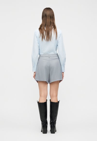 Γυναίκα με μακριά καστανά μαλλιά στέκεται με την πλάτη στραμμένη φορώντας ένα ανοιχτό μπλε πουκάμισο με μακριά μανίκια, γκρι σορτς και μαύρες μπότες μέχρι το γόνατο.