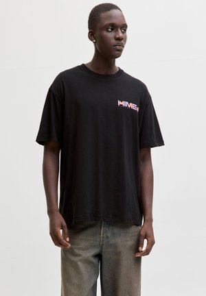 Νεαρός άνδρας που φοράει μαύρο oversized μπλουζάκι με πορτοκαλί γραφικό κείμενο στο στήθος και φθαρμένο μπλε τζιν, στέκεται μπροστά σε απλό λευκό φόντο.