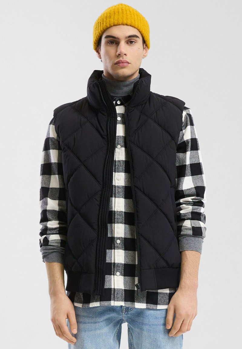 Schwarze gesteppte Weste mit hohem Kragen über einem schwarz-weißen karierten Hemd, kombiniert mit hellblauen Jeans und einer gelben Strickmütze.