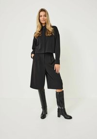 Haut noir court à manches longues associé à un short large taille haute, complété par des bottes noires brillantes jusqu'aux genoux. Tissu au toucher lisse.