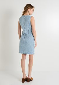 Robe en denim à silhouette ajustée, de couleur bleu clair, sans manches, avec un ourlet effiloché et un détail de fermeture éclair au dos. Accompagnée de sandales à talons marron.