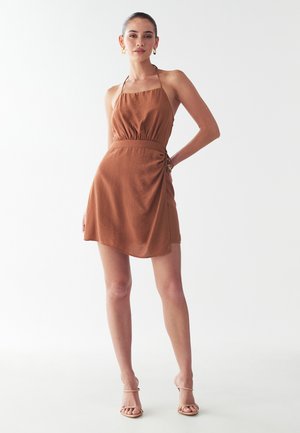 Abito halter marrone in tessuto testurizzato, con vita elastica e dettagli arricciati sui fianchi. Abbinato a sandali nudi con cinturini.