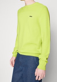 Man som bär en limegrön långärmad tröja med "BOSS"-logotyp på bröstet och blå strukturerade byxor mot en enkel ljus bakgrund.