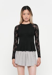 Haut noir à manches longues avec des manches en dentelle, ourlet peplum, associé à une jupe plissée gris clair. Design simple avec des textures contrastées.