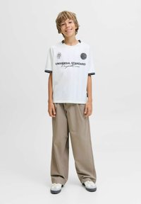 Ragazzino sorridente in piedi, indossa una maglietta bianca con grafica, pantaloni beige larghi e sneakers bianche su uno sfondo grigio uniforme.
