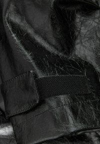 Juoda, struktūruota dirbtinė oda, pasižyminti velcro juosta ir siuvimo detalėmis, rodanti blizgantį paviršių ir šiek tiek raukšlių medžiagoje.