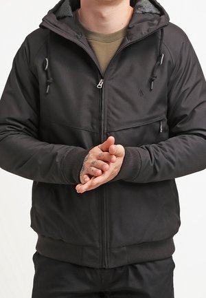 Homme portant une veste noire à fermeture éclair avec capuche et poignets côtelés, mains jointes montrant des tatouages sur les doigts sur un fond uni.