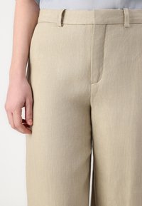 Beige linbukser med rett ben-design, med flat front, sidelommer og beltehemper. Strukturert stoff er synlig.