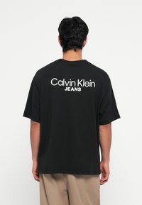 Crna pamučna majica koja ima slobodan kroj i kratke rukave, s natpisom "Calvin Klein JEANS" u bijeloj boji na leđima.