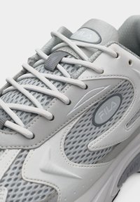 Ανοιχτό γκρι και λευκό αθλητικό παπούτσι FILA με πάνελ από δίχτυ, λευκά κορδόνια και λογότυπα με το εμπορικό σήμα στη γλώσσα και στην πλευρά.