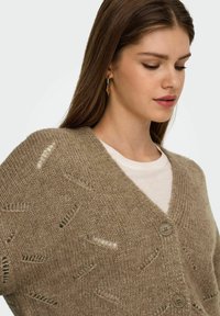 Beige strik cardigan med V-hals, der har teksturerede mønstre og to knapper foran. Bæres over en lyse farvet t-shirt.