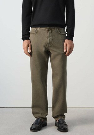 Mann trägt olivgrüne gerade Hose, schwarzes langärmeliges Hemd und schwarze Lederschuhe, steht vor einem einfarbigen Hintergrund.