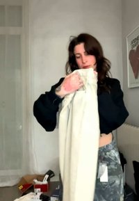 Žena s tmavými vlasmi drží a skúma pár svetlých nohavíc v miestnosti s krabicami na oblečenie a zarámovaným obrazom srdca.