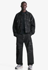 Sort denimjakke med hvide syninger, parret med sorte cargo-bukser med lommer. Model iført grå sneakers står på en hvid baggrund.