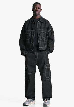 Black denim jacket with white stitching, paired with black cargo pants featuring pockets. Model wearing gray sneakers stands on a white background.