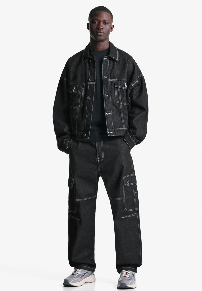 Sort denimjakke med hvide syninger, parret med sorte cargo-bukser med lommer. Model iført grå sneakers står på en hvid baggrund.