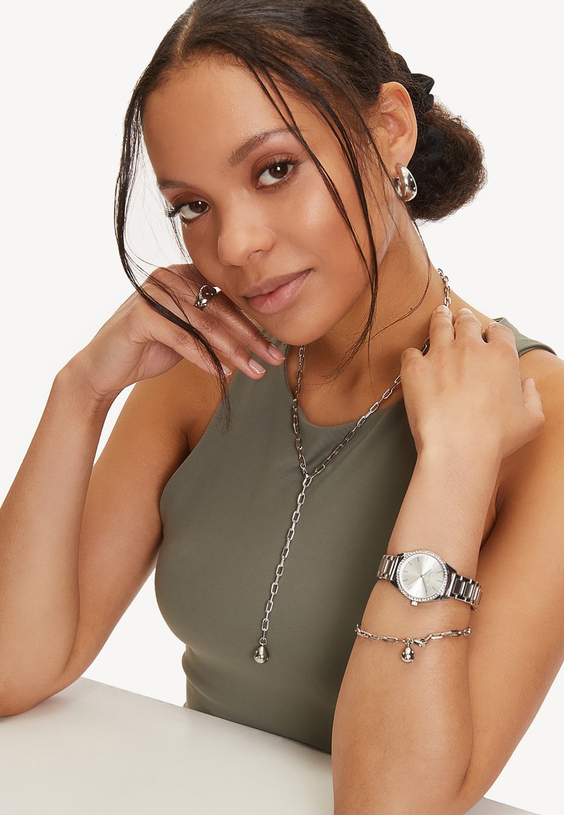 Silberne Uhr mit rundem Ziffernblatt, Gliederarmband und Halskette mit Anhänger. Das Modell trägt ein enges olivgrünes Tanktop, das den Schmuck zur Schau stellt.