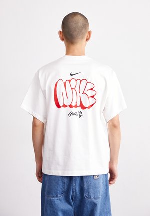T-shirt en coton blanc arborant un texte "NIKE" audacieux de style graffiti rouge au dos, avec un logo Nike noir au-dessus et "Since '12" en dessous.