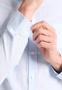 Ljusblå linneskjorta med knappar, spetsig krage och enfärgad manschett med en knapp. Har en bröstficka och en slät textur.