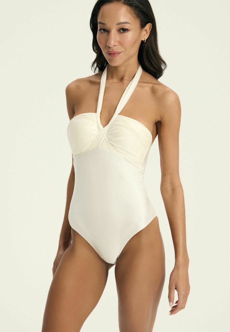 Jednoczęściowy strój kąpielowy w jasnym kremowym kolorze, z dekoltem na halter, marszczonym korpusem, gładką teksturą i wysokimi wycięciami na nogawkach.