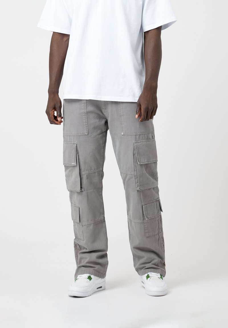 Homme portant un pantalon cargo gris clair avec plusieurs poches, des baskets blanches et un t-shirt blanc uni à manches courtes sur un fond gris uni.
