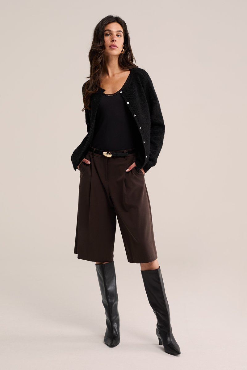 Frau trägt schwarzen Cardigan, schwarzes Top, dunkelbraune knieumspielende weite Hose, schwarzen Gürtel und schwarze kniehohe Stiefel und steht mit den Händen in den Taschen.