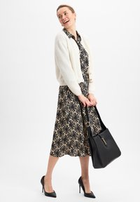 Weiße flauschige Strickjacke über einem schwarz-blumig gemusterten Kleid, kombiniert mit schwarzen hohen Schuhen und einer schwarzen Lederhandtasche.