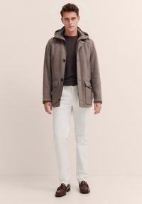 Grauer Wollmischmantel mit Kapuze, knöpfbarem Vorderteil und zwei Fronttaschen, getragen über einem dunklen Pullover und weißen Hosen, kombiniert mit braunen Schuhen.